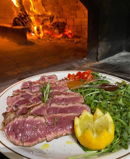 Tagliata al sangue con rosmarino, rucola, pomodorini e limone su piatto bianco davanti al forno a legna acceso